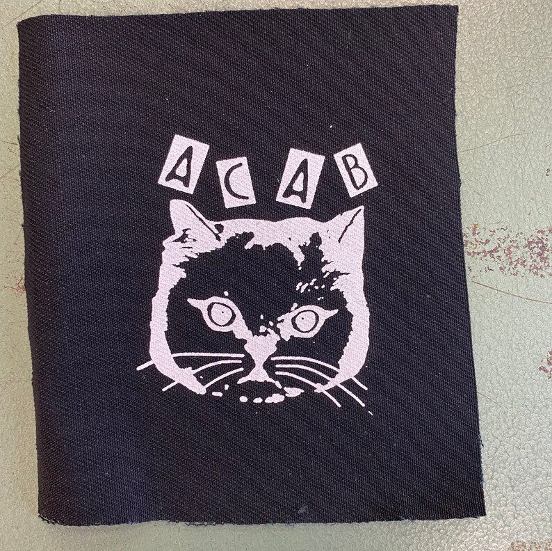 K&ouml;nnte beinhalten: Schwarz-wei&szlig;es B&uuml;geleisen-Patch mit einem Katzenkopf und den Buchstaben "ACAB" dar&uuml;ber.