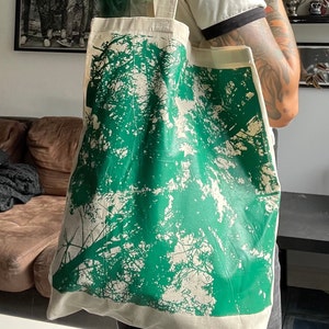 May include: A white canvas tote bag with a green tree silhouette print. The bag has a long handle and is being carried by a person.