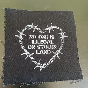 May include: Black fabric patch with white text and a barbed wire heart design. The text reads "NO ONE IS ILLEGAL ON STOLEN LAND". The patch has a raw edge.