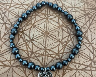May include: A dark grey beaded bracelet with a silver heart pendant. The bracelet is made of small, round beads and features a decorative heart charm. The bracelet is displayed on a wooden surface with a geometric pattern.