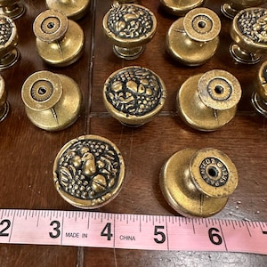 May include: A collection of round brass-toned metal cabinet knobs featuring a raised fruit motif on the face. The back of each knob is stamped with the text 1335 PATENTED. A pink measuring tape is visible, showing the knobs are approximately 1.5 inches.