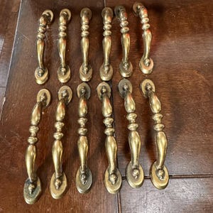 May include: A set of twelve brass cabinet pulls arranged in two rows of six. Each pull features a curved, elongated handle with a central bulbous section and rounded mounting plates at both ends, showcasing a polished metallic gold finish.