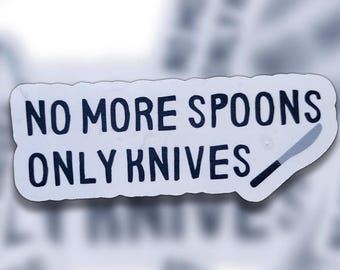 以下が含まれることがあります： 「NO MORE SPOONS ONLY KNIVES」の黒い文字とナイフのイラストが描かれた白いステッカー。背景は少しぼやけています。