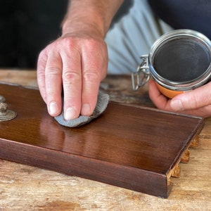 May include: A person's hand rubs a dark gray polishing cloth on a dark brown wooden surface. A glass jar with a metal lid containing a dark brown substance is in the background.