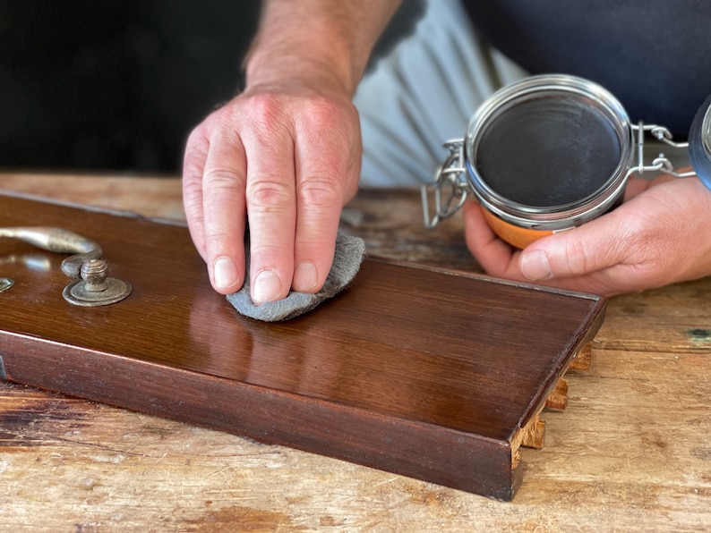 May include: A person's hand rubs a dark gray polishing cloth on a dark brown wooden surface. A glass jar with a metal lid containing a dark brown substance is in the background.