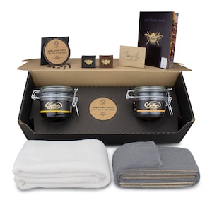 May include: A black gift box with a brown lid, containing two glass jars of honey, two small gold bee-shaped charms, a thank you card, a small bag of sunflower seeds, and two towels, one white and one gray.