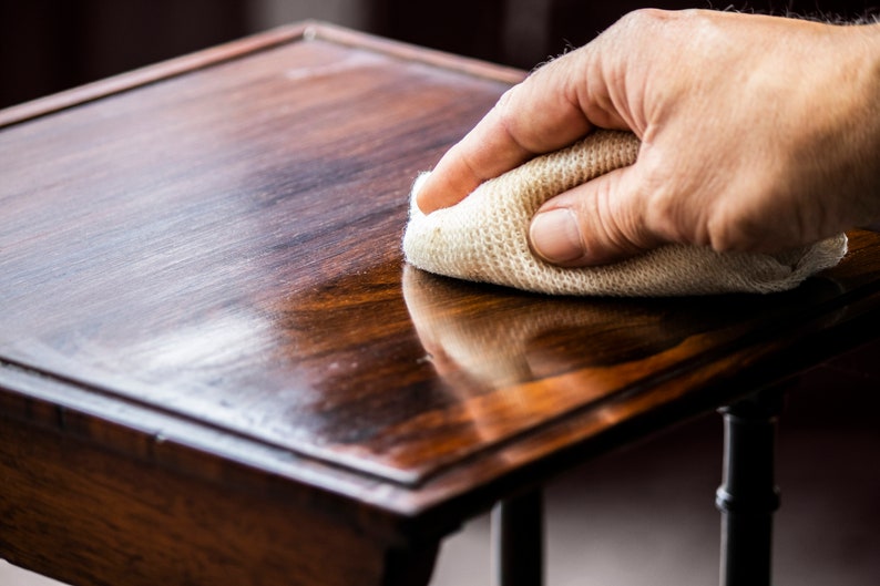 May include: A hand rubs a white cloth over a dark wood table top, polishing the surface to a shine.