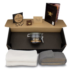 Puede incluir: Una caja de cartón marrón con interior negro. La caja contiene un tarro de vidrio de betún negro, dos paños, una pequeña caja de madera, una tarjeta que dice "Gracias", una tarjeta que dice "Semillas de girasol para ti y las abejas", y un folleto que dice "No mires dentro".