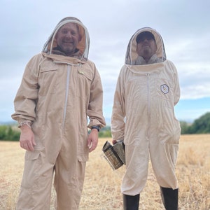 May include: Two people wearing beige beekeeping suits with veils and protective headgear stand in a field. One person is holding a smoker.