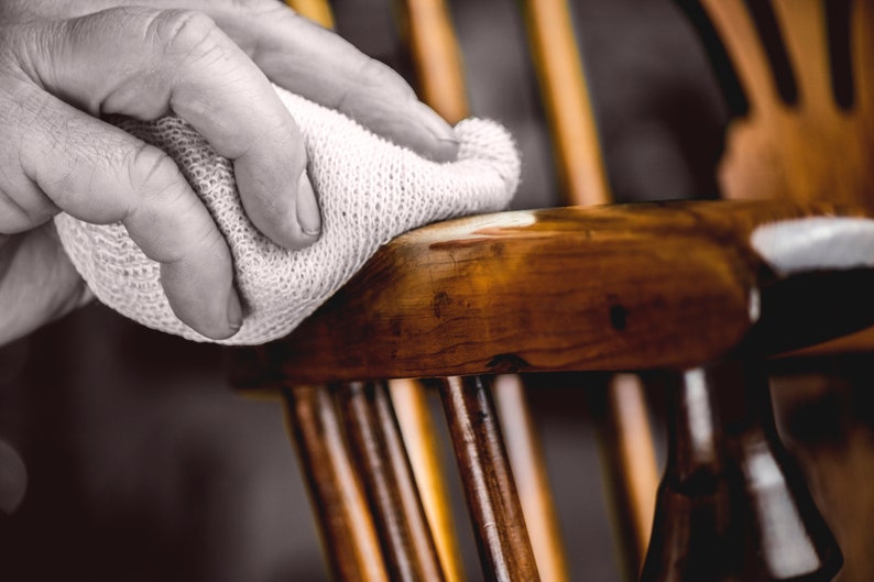 May include: A person's hand is using a white cloth to polish a wooden chair. The chair has a dark brown finish and is made of wood.
