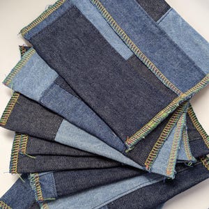 Op de afbeelding: Een stapel denim onderzetters in verschillende blauwtinten. Elke onderzetter is vierkant met een patchwork ontwerp en een kleurrijke gestikte rand. De onderzetters zijn in een waaier vorm gerangschikt, waarbij de verschillende denim tinten worden getoond.
