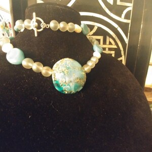 May include: A turquoise and gold necklace with a large, round pendant. The pendant is made of glass and has a marbled effect. The necklace is made of white and turquoise beads.
