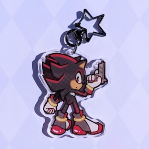 May include: A clear acrylic keychain featuring a cartoon character with black and red accents. The character is holding a gun and has a star-shaped clasp. The keychain is a collectible item.