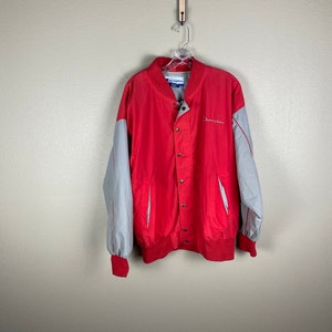 May include: A red and grey Champion baseball jacket with a snap button closure. The jacket has a grey stripe down the sleeves and the Champion logo on the right side.