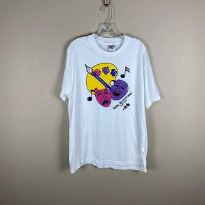 May include: White t-shirt with a colorful graphic design featuring a paint palette, paintbrush, musical notes, and two theatrical masks. The text "Girl Scouting and the Arts" is printed on the shirt.