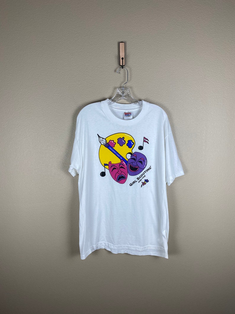 May include: White t-shirt with a colorful graphic design featuring a paint palette, paintbrush, musical notes, and two theatrical masks. The text "Girl Scouting and the Arts" is printed on the shirt.