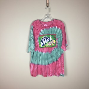 May include: A tie-dye t-shirt in shades of pink and teal, hanging on a hanger. The shirt features a Sierra Mist Cranberry Splash logo on the front. The shirt is a short-sleeved, casual style.