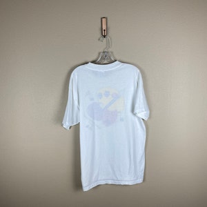 May include: White t-shirt with a colorful graphic of a cartoon character on the back. The character is wearing a blue hat and holding a paintbrush.