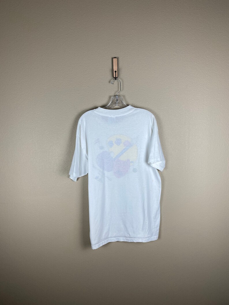 May include: White t-shirt with a colorful graphic of a cartoon character on the back. The character is wearing a blue hat and holding a paintbrush.