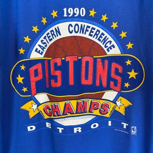 Puede incluir: Una camiseta azul con un gr&aacute;fico blanco, rojo y amarillo del logotipo de los Detroit Pistons 1990 Eastern Conference Champions. El logotipo presenta un bal&oacute;n de baloncesto con la palabra "PISTONS" en letras rojas y la palabra "CHAMPS" en letras amarillas. El texto "DETROIT" est&aacute; impreso debajo del logotipo.