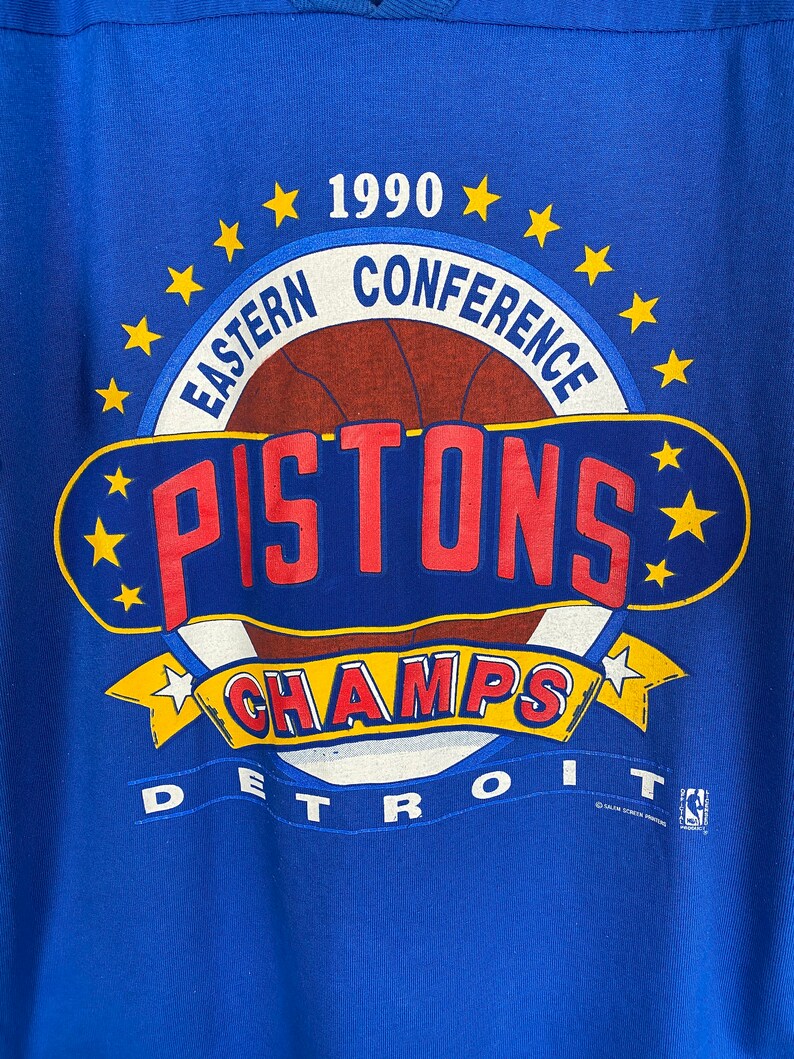 Puede incluir: Una camiseta azul con un gr&aacute;fico blanco, rojo y amarillo del logotipo de los Detroit Pistons 1990 Eastern Conference Champions. El logotipo presenta un bal&oacute;n de baloncesto con la palabra "PISTONS" en letras rojas y la palabra "CHAMPS" en letras amarillas. El texto "DETROIT" est&aacute; impreso debajo del logotipo.