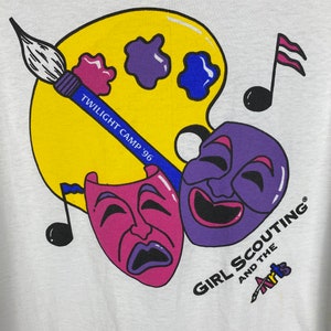 May include: A white t-shirt with a colorful graphic design. The design features a paint palette, paintbrushes, musical notes, and two theatrical masks. The text "Twilight Camp '96" and "Girl Scouting and the Arts" are included in the design.