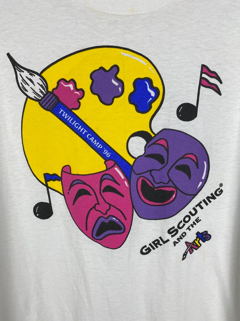 May include: A white t-shirt with a colorful graphic design. The design features a paint palette, paintbrushes, musical notes, and two theatrical masks. The text "Twilight Camp '96" and "Girl Scouting and the Arts" are included in the design.