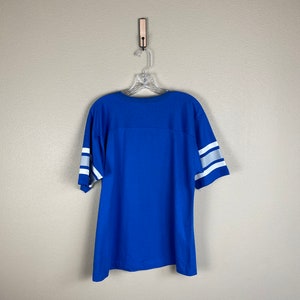 Puede incluir: Una camiseta azul real con rayas blancas en las mangas. La camiseta est&aacute; colgada en una percha de madera contra una pared gris claro.