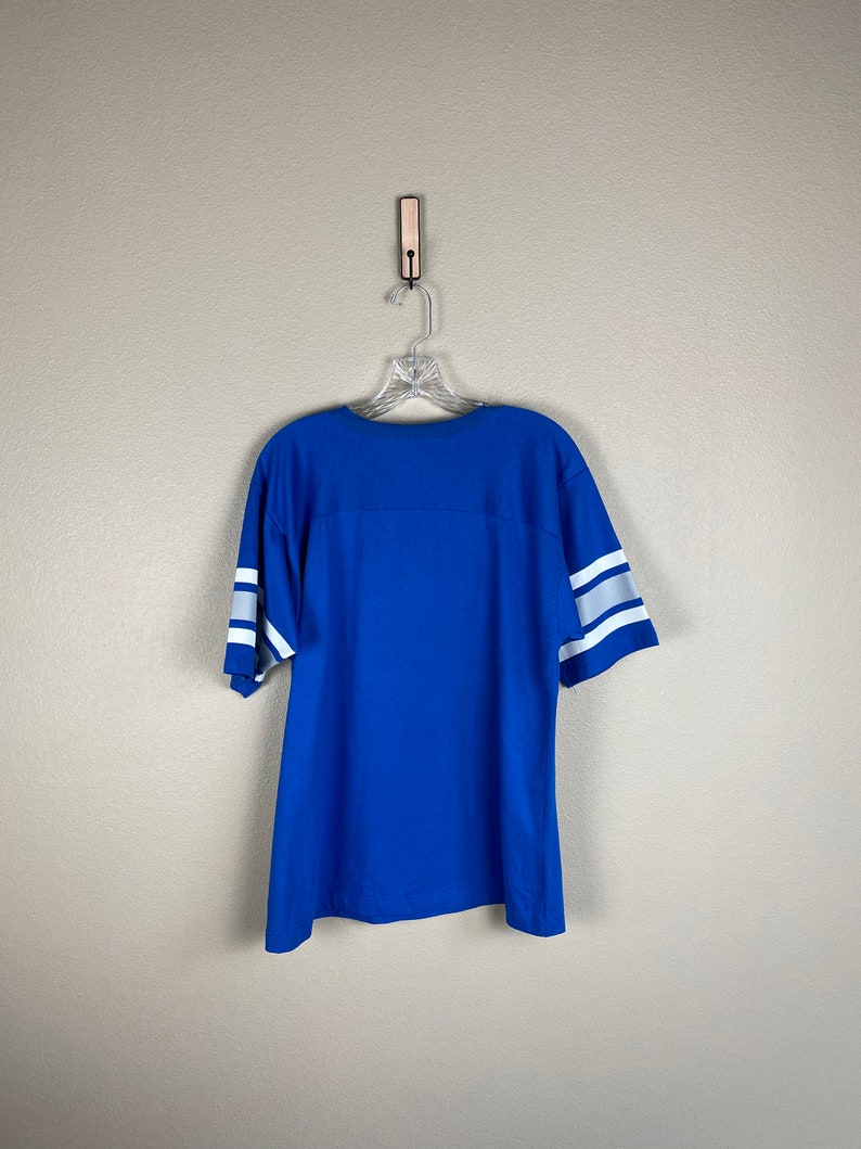 Puede incluir: Una camiseta azul real con rayas blancas en las mangas. La camiseta est&aacute; colgada en una percha de madera contra una pared gris claro.