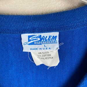 Puede incluir: Una camiseta azul con una etiqueta blanca que dice "Salem Sportswear" y "Made in U.S.A." La etiqueta tambi&eacute;n incluye la informaci&oacute;n "RN 52325", "50% Algod&oacute;n" y "50% Poli&eacute;ster".