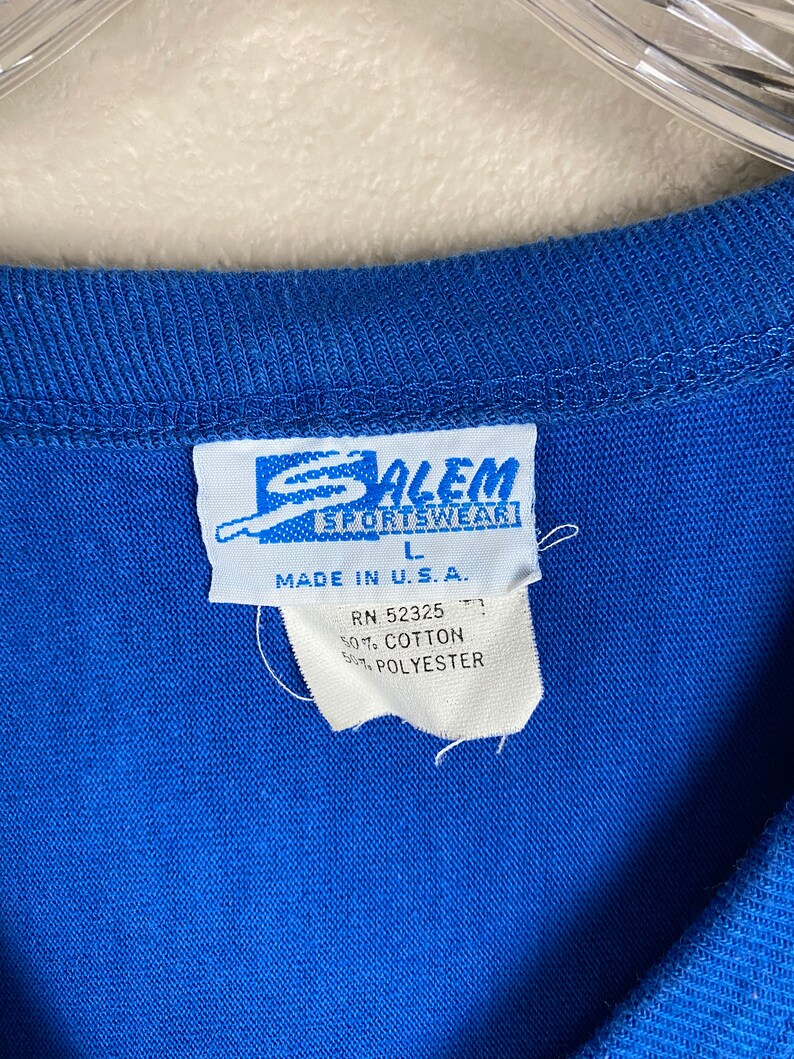 Puede incluir: Una camiseta azul con una etiqueta blanca que dice "Salem Sportswear" y "Made in U.S.A." La etiqueta tambi&eacute;n incluye la informaci&oacute;n "RN 52325", "50% Algod&oacute;n" y "50% Poli&eacute;ster".