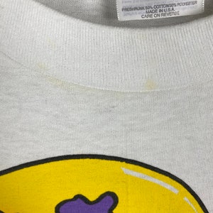 May include: White t-shirt with a yellow smiley face graphic. The smiley face has purple, pink, and blue spots. The shirt tag says "Heavyweight 50/50 Adult L (42-44) Preshrunk 50% Cotton/50% Polyester Made in USA Care on Reverse"