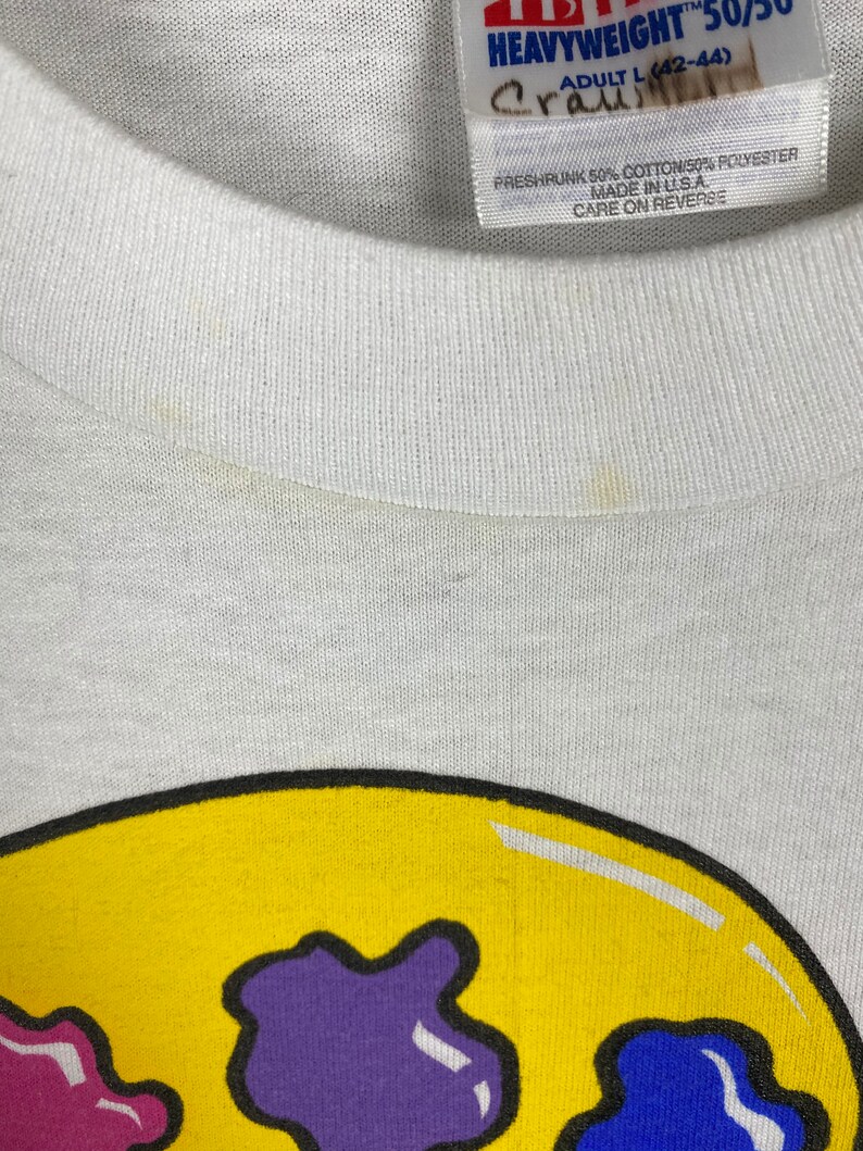 May include: White t-shirt with a yellow smiley face graphic. The smiley face has purple, pink, and blue spots. The shirt tag says "Heavyweight 50/50 Adult L (42-44) Preshrunk 50% Cotton/50% Polyester Made in USA Care on Reverse"