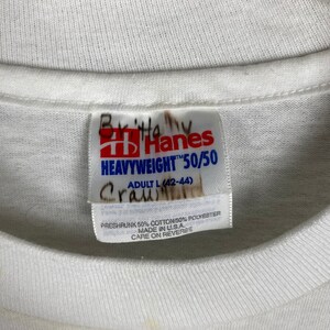 May include: White Hanes Heavyweight 50/50 T-shirt tag with size label Adult L (42-44). The tag is made of white fabric with blue text and a red Hanes logo. The tag is preshrunk 50% cotton and 50% polyester, made in the USA.