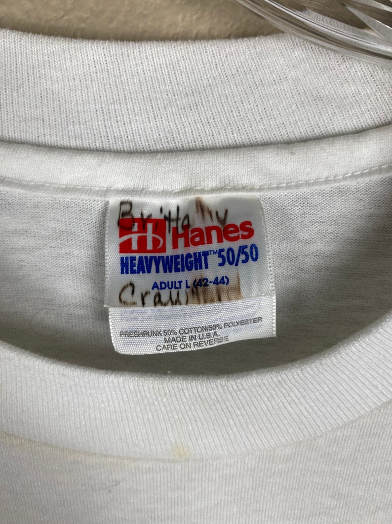 May include: White Hanes Heavyweight 50/50 T-shirt tag with size label Adult L (42-44). The tag is made of white fabric with blue text and a red Hanes logo. The tag is preshrunk 50% cotton and 50% polyester, made in the USA.