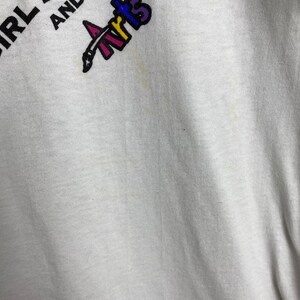 May include: White t-shirt with the text "Girl Scouts and the Arts" in black and purple lettering. A colorful paintbrush logo is above the text.
