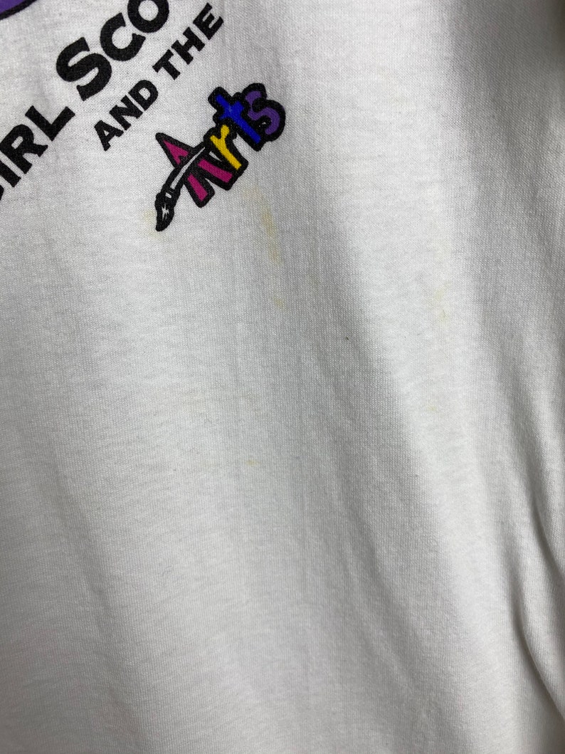 May include: White t-shirt with the text "Girl Scouts and the Arts" in black and purple lettering. A colorful paintbrush logo is above the text.
