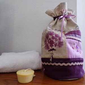 Peut inclure: Un sac en tissu de couleur lavande avec un motif de trou de serrure et un ruban violet. Le sac est fait de tissu patchwork avec un motif floral et une bordure en dentelle. Le sac est posé sur une surface en bois à côté d'une serviette blanche et d'une petite bougie.