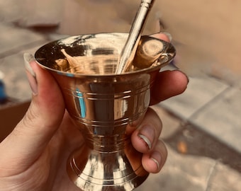 May include: A gold metal mortar and pestle set. The mortar is a small, round bowl with a flared lip. The pestle is a long, thin rod with a rounded end.