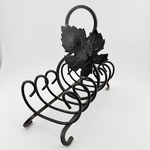 May include: A black metal napkin holder featuring a curved, scrollwork design and a decorative grape leaf accent. The structure is composed of thin, dark metal rods formed into a series of loops, supported by four small, curled metal feet at the base.