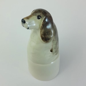 Puede incluir: Un tapón de botella de cerámica con forma de perro pintado en marrón y blanco. El perro está sentado con la cabeza inclinada hacia un lado.