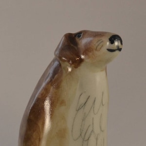 May include: A ceramic figurine of a brown and white dog with a white base. The dog is looking up and has a black nose. The figurine is signed "Sue Lee" on the side.