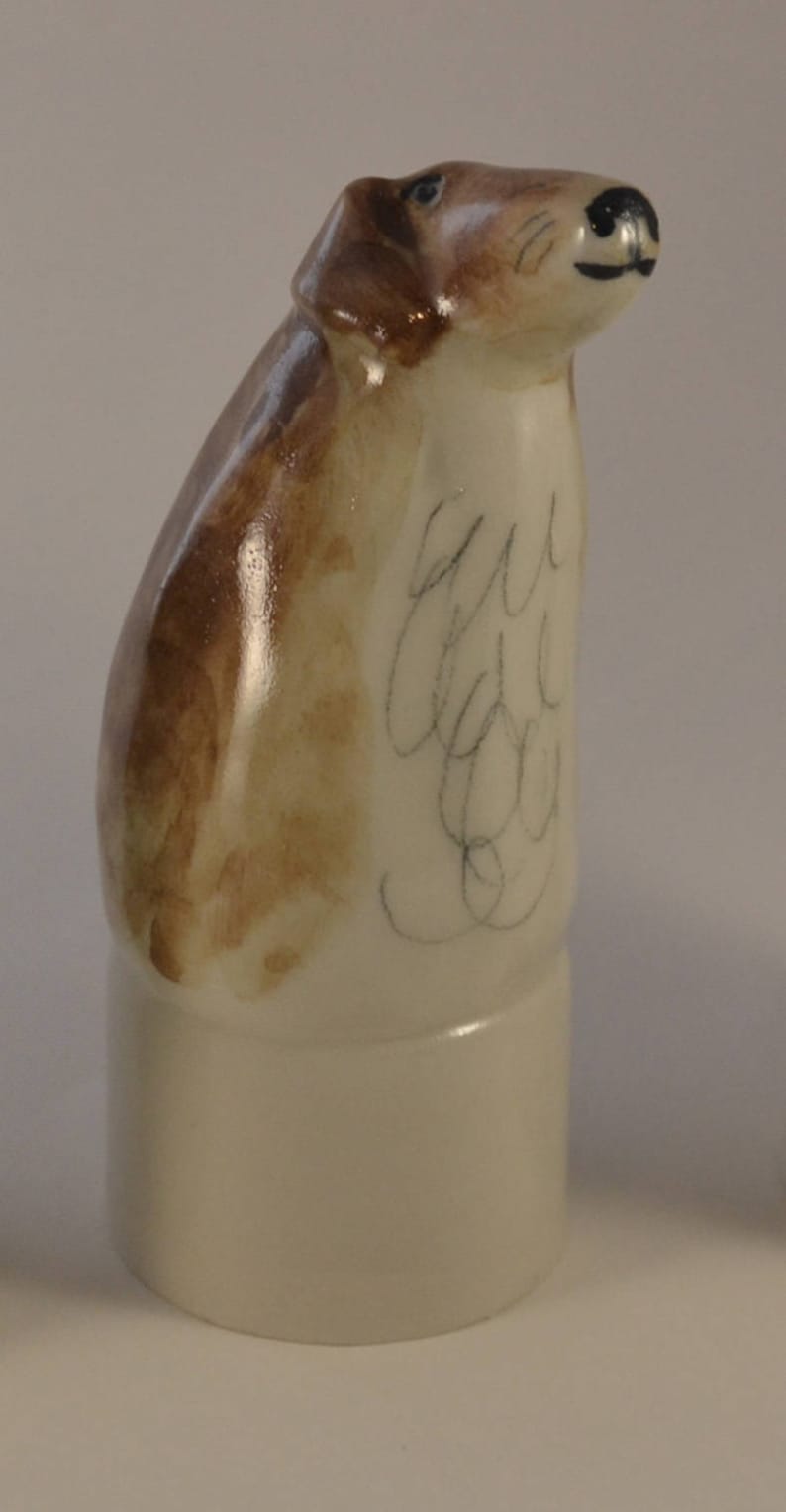 May include: A ceramic figurine of a brown and white dog with a white base. The dog is looking up and has a black nose. The figurine is signed "Sue Lee" on the side.