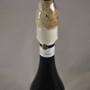 May include: A ceramic bottle stopper shaped like a dog, with brown and white markings. The stopper is on top of a wine bottle with a black label that says "First Selection".