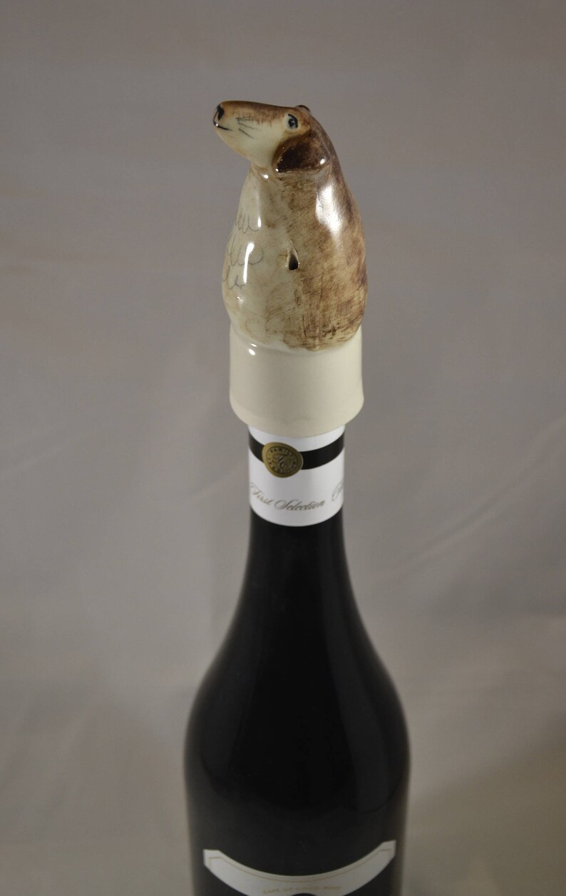 May include: A ceramic bottle stopper shaped like a dog, with brown and white markings. The stopper is on top of a wine bottle with a black label that says "First Selection".