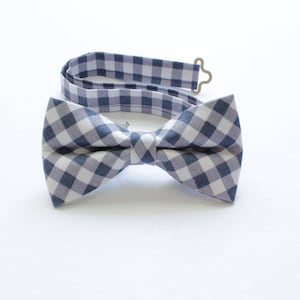 May include: A blue and white gingham bow tie with an adjustable strap and metal clasp. The bow tie is made of fabric and is displayed on a white background. The bow tie is a classic accessory.