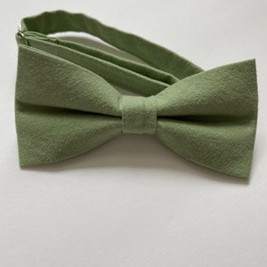 May include: A sage green bow tie made of textured fabric. The bow tie is pre-tied and has an adjustable strap. The fabric has a subtle, natural texture. The bow tie is a classic accessory for formal or semi-formal attire.