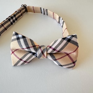 May include: A beige plaid bow tie with black and burgundy accents. The bow tie is pre-tied and has an adjustable strap. The pattern is a classic tartan design, suitable for formal or semi-formal occasions.