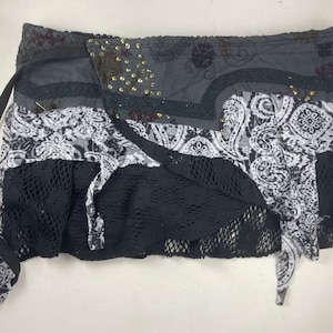 Op de afbeelding: Een zwarte en witte geprinte wikkelrok met een zwarte mesh overlay en een zwarte strik. De rok heeft een bohemian stijl en is versierd met goudkleurige versieringen.