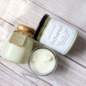 May include: Two glass jars with cork lids filled with white soy wax candles. The labels on the jars read "Enchanted" and "Peach - Lily - Cedar". The candles are handcrafted and have a burn time of approximately 50 hours.